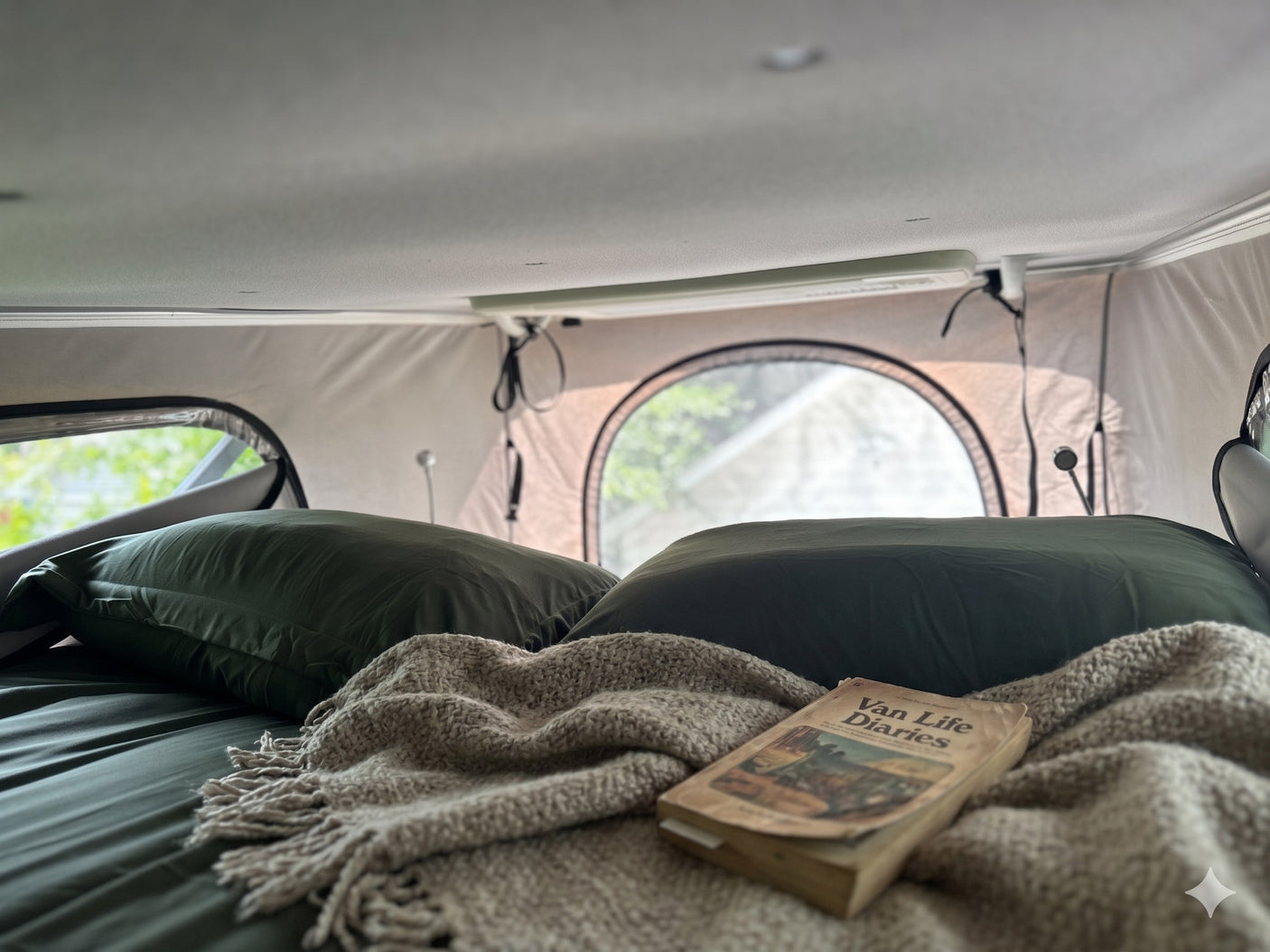 Inside a cozy RV rental: bed with green pillows, a textured blanket, and "Van Life Diaries" book.