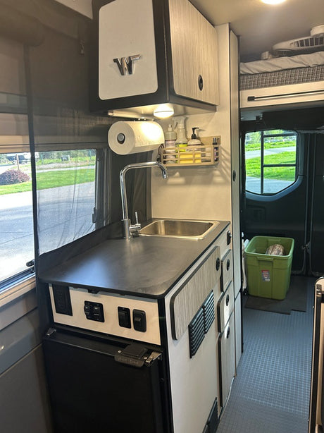 Interior of an RV rental showing a compact galley kitchen with sink, faucet, small fridge, and overhead cabinets. Ideal for travel.