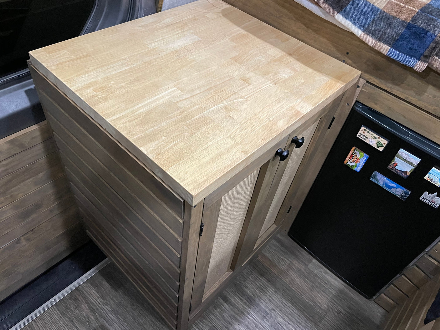 RV rental interior showing a light wood block countertop on a slatted cabinet, next to a black mini-fridge with magnets.