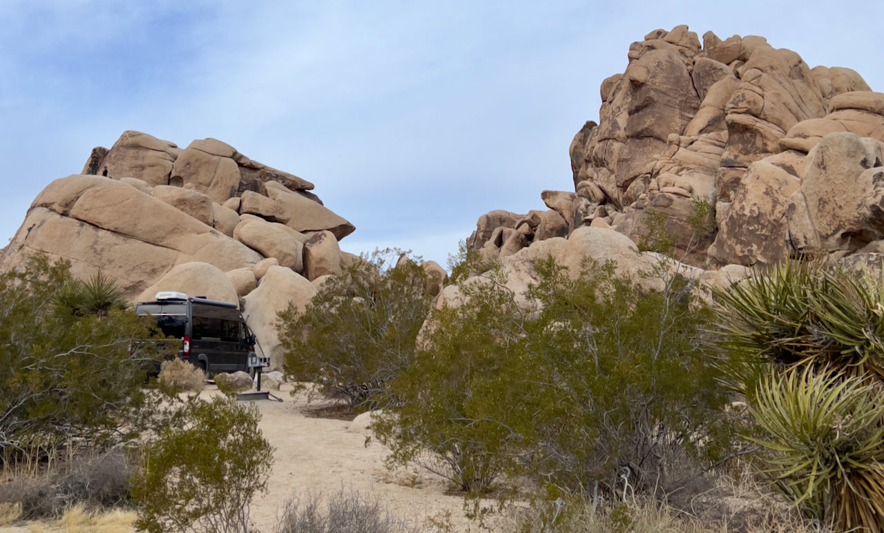 RV rental camper van parked at a scenic desert campsite, nestled among massive rock formations and native plants under a wide sky.