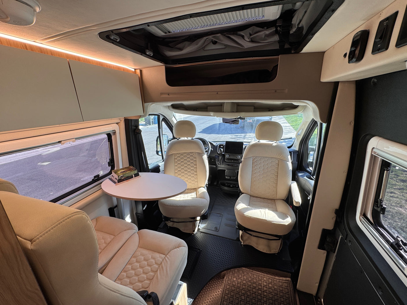 Modern rv rental interior showing cream swivel front seats, a dining table, and illuminated overhead storage. Spacious camper van.