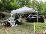 Camping setup featuring a teardrop rv rental with an open kitchen, SUV, and shade canopy in a wooded area.