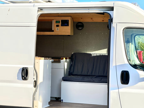 White camper van RV rental interior: custom control panel, kitchenette, and bench seating visible through open side door.