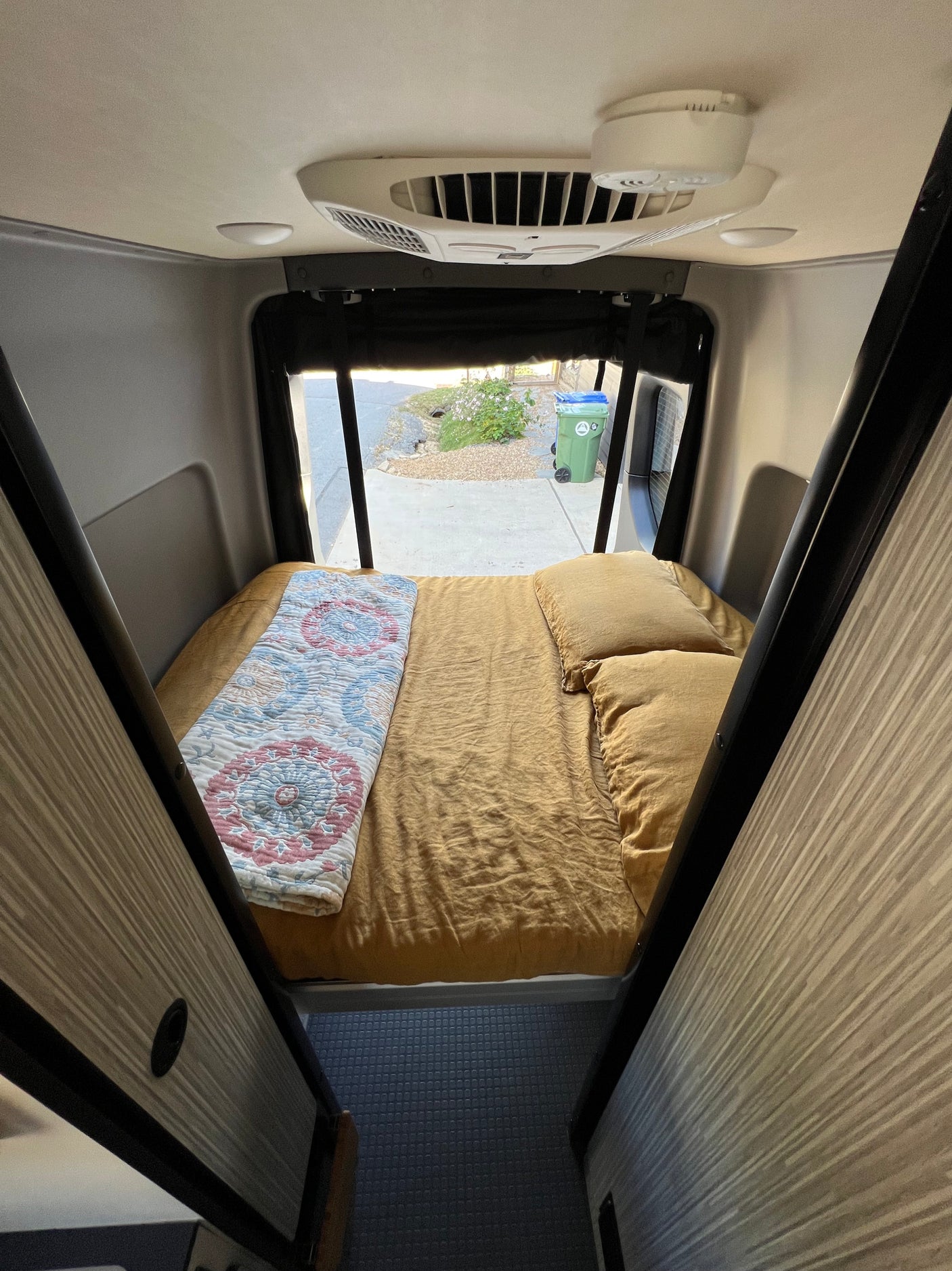 Inside a cozy RV rental: a comfortable bed, overhead AC, and a view through the open rear. A compact mobile living space.