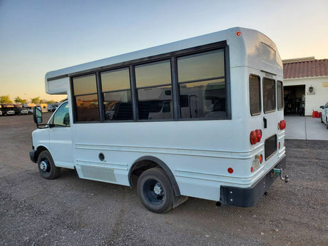 White shuttle bus, potential RV rental conversion, parked on gravel. Tinted windows, red tail lights, and tow hitch visible.