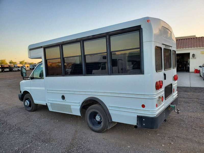 White shuttle bus, potential RV rental conversion, parked on gravel. Tinted windows, red tail lights, and tow hitch visible. — photo 5