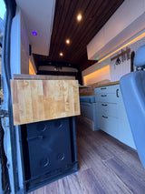 RV rental kitchen setup featuring a pull-out countertop, hidden fridge, custom cabinets, wood ceiling, and comfy seating.