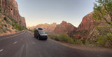 A red RV rental van is parked on a scenic road in Zion National Park, surrounded by towering red rock mountains.