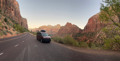 A red RV rental van is parked on a scenic road in Zion National Park, surrounded by towering red rock mountains.