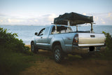 Silver Toyota Tacoma pickup with a rooftop tent, parked at a sandy beach with ocean views. Ideal for RV rental style beach camping.