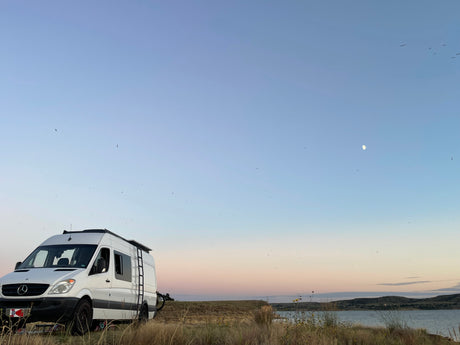 Enjoy van life adventures with an RV rental! Sunset scene by a lake with birds and moon.