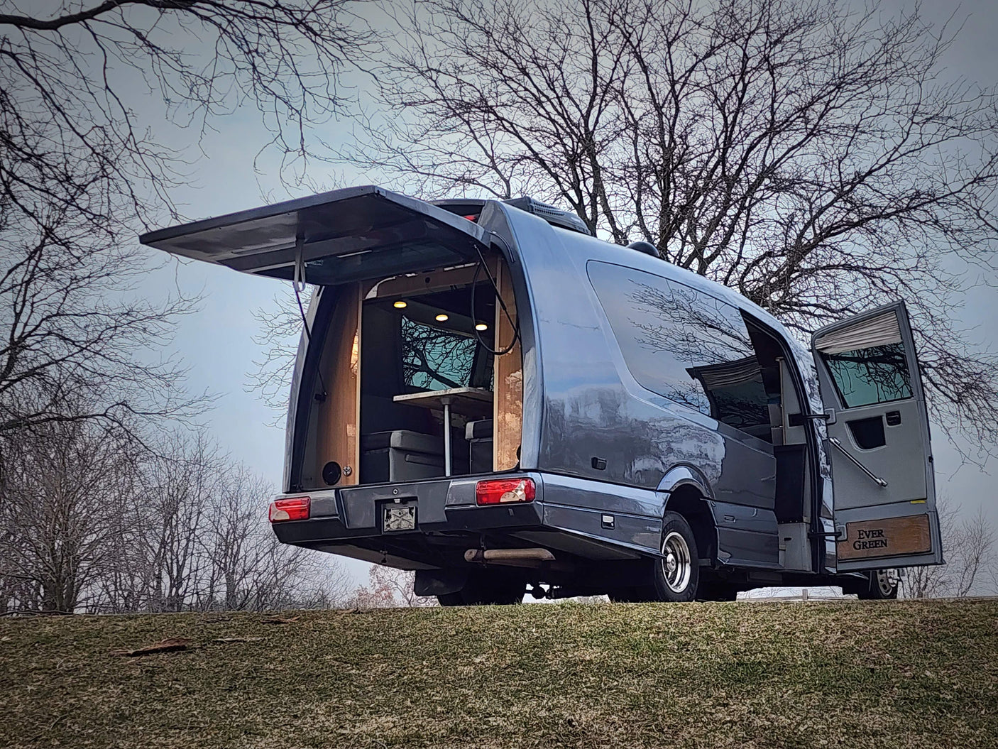 This gray custom camper van, perfect for rv rental, reveals its cozy, lit interior with open rear hatch & side door on a grassy hill.