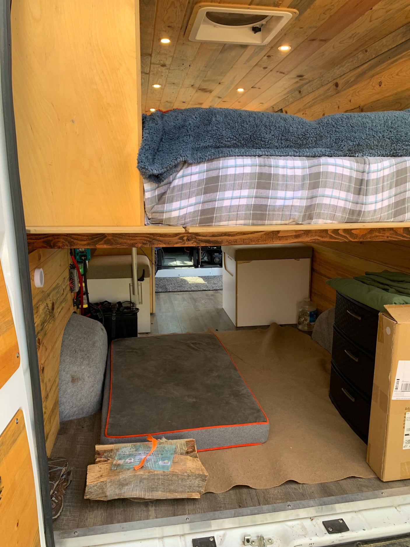 Inside a comfy RV rental: raised bed, wood-paneled walls/ceiling, lights, and floor space with a dog bed. Ready for van life!