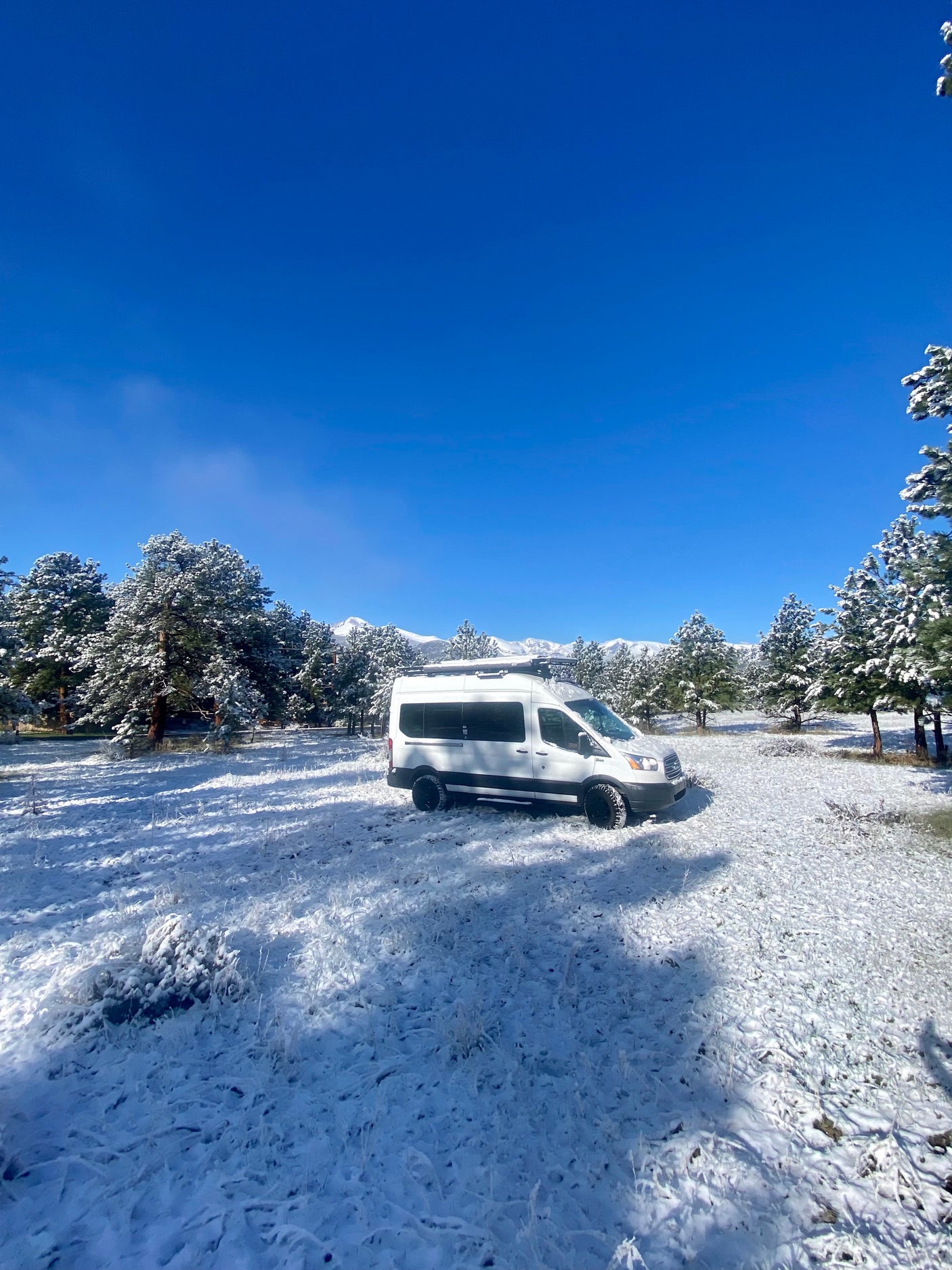 White RV rental camper van parked in a snowy landscape with frosted pine trees and distant snow-capped mountains.
