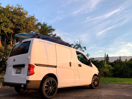 White Nissan van with surfboard on roof, a perfect RV rental for island adventures in a tropical setting.