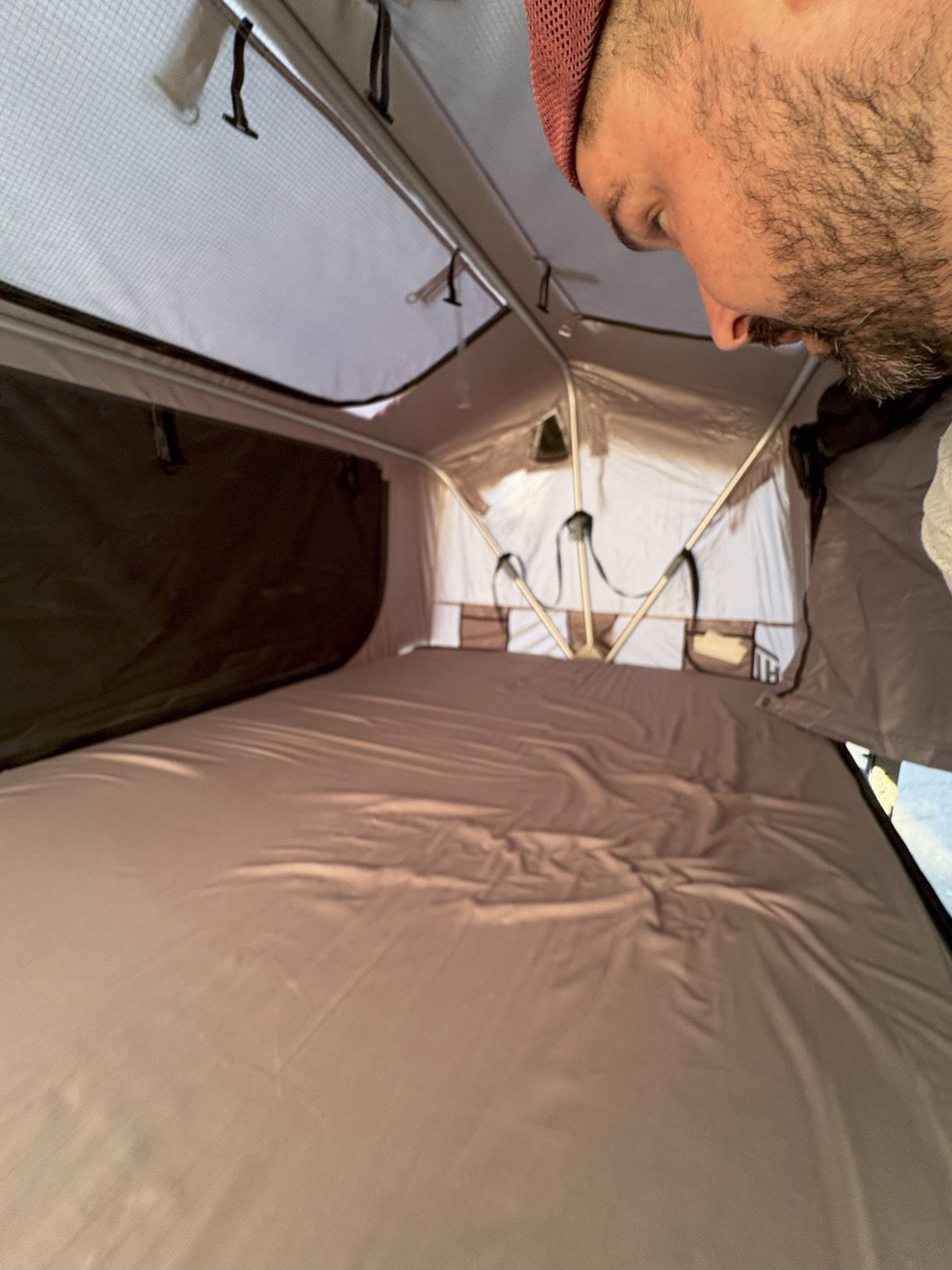 Inside an RV rental's pop-up tent, showing a comfortable sleeping area with a neatly made bed and storage pockets. A man peers in from above.