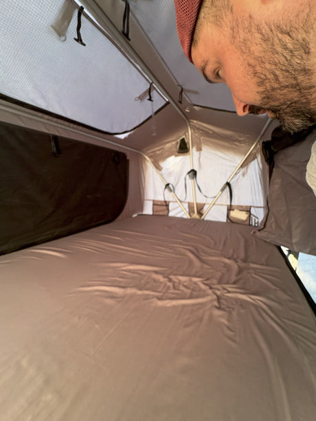 Inside an RV rental's pop-up tent, showing a comfortable sleeping area with a neatly made bed and storage pockets. A man peers in from above.