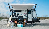 An RV rental parked on a sandy beach. Two women relax in chairs by the white travel trailer under an awning, with a surfboard & cooler.