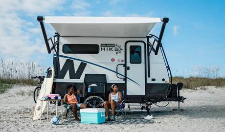 An RV rental parked on a sandy beach. Two women relax in chairs by the white travel trailer under an awning, with a surfboard & cooler.