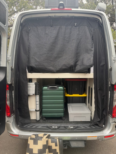 Rear of an rv rental van with doors open, showing organized interior storage: bed, luggage, bins, and a mesh screen.