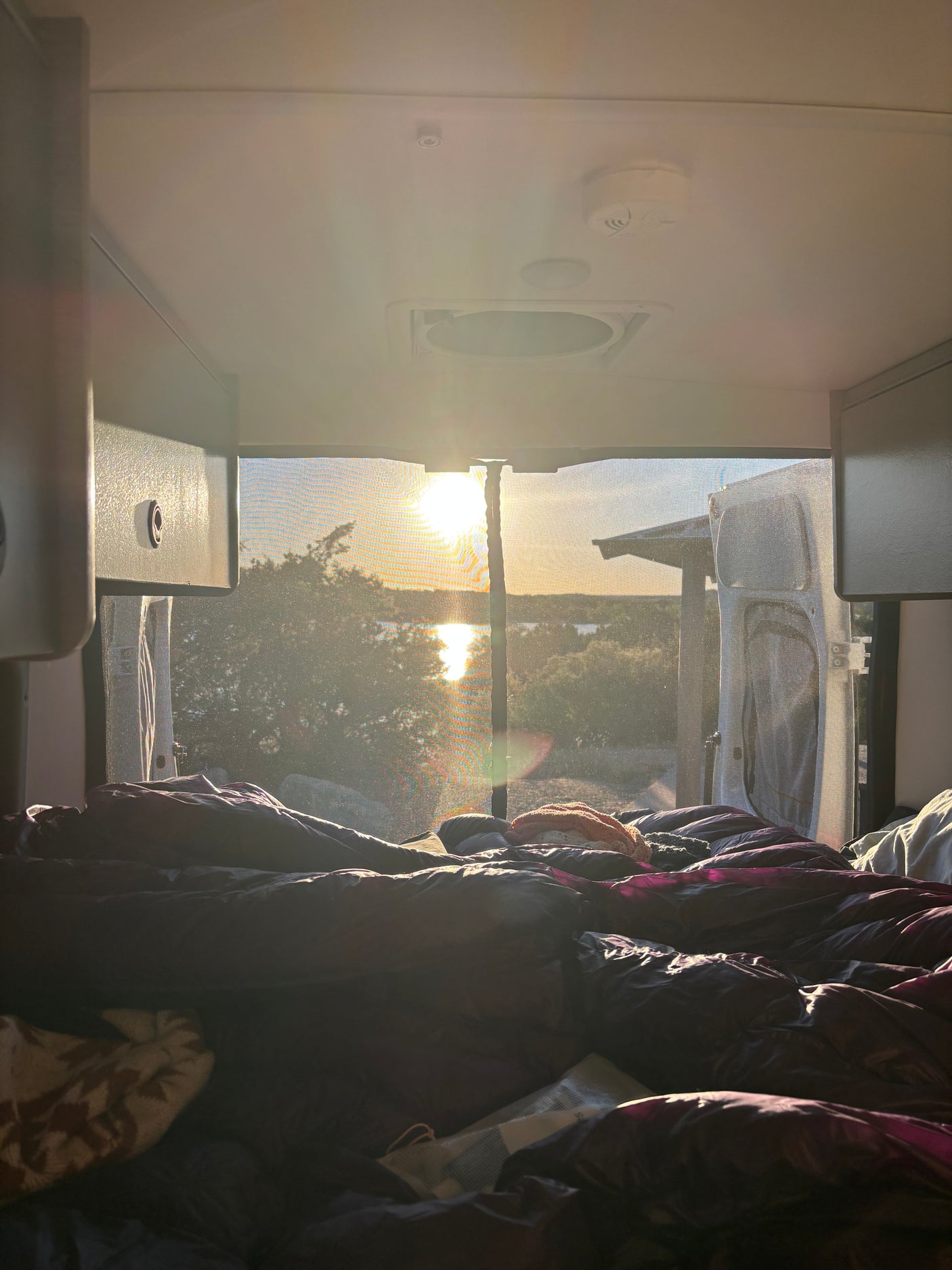 RV rental bedroom with a view: golden sun reflecting on water through a screened window, seen from a cozy bed.