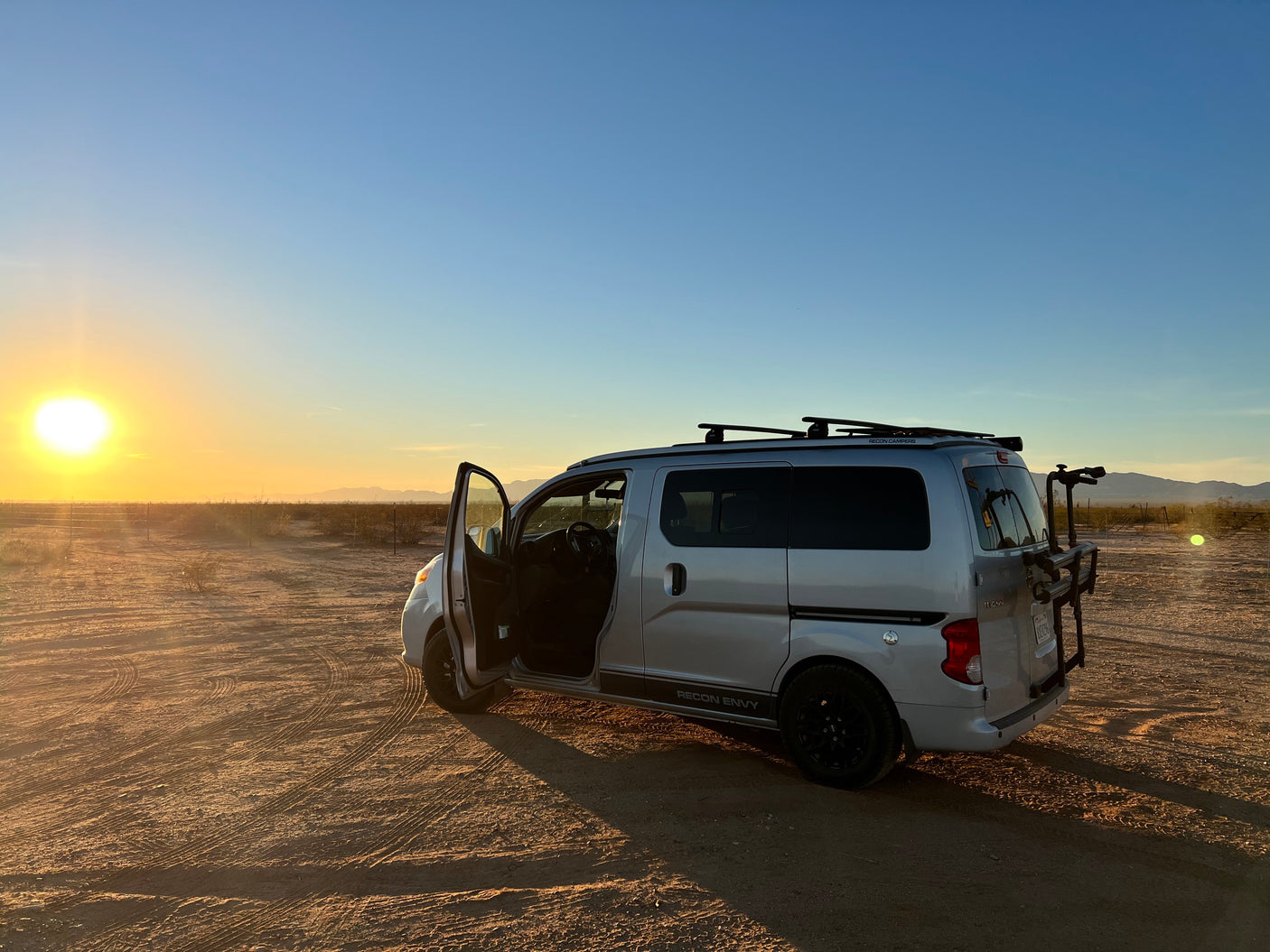 A silver Recon Camper van (RV rental) is parked in a desert at sunset, driver's door open, with a bike rack on the rear.