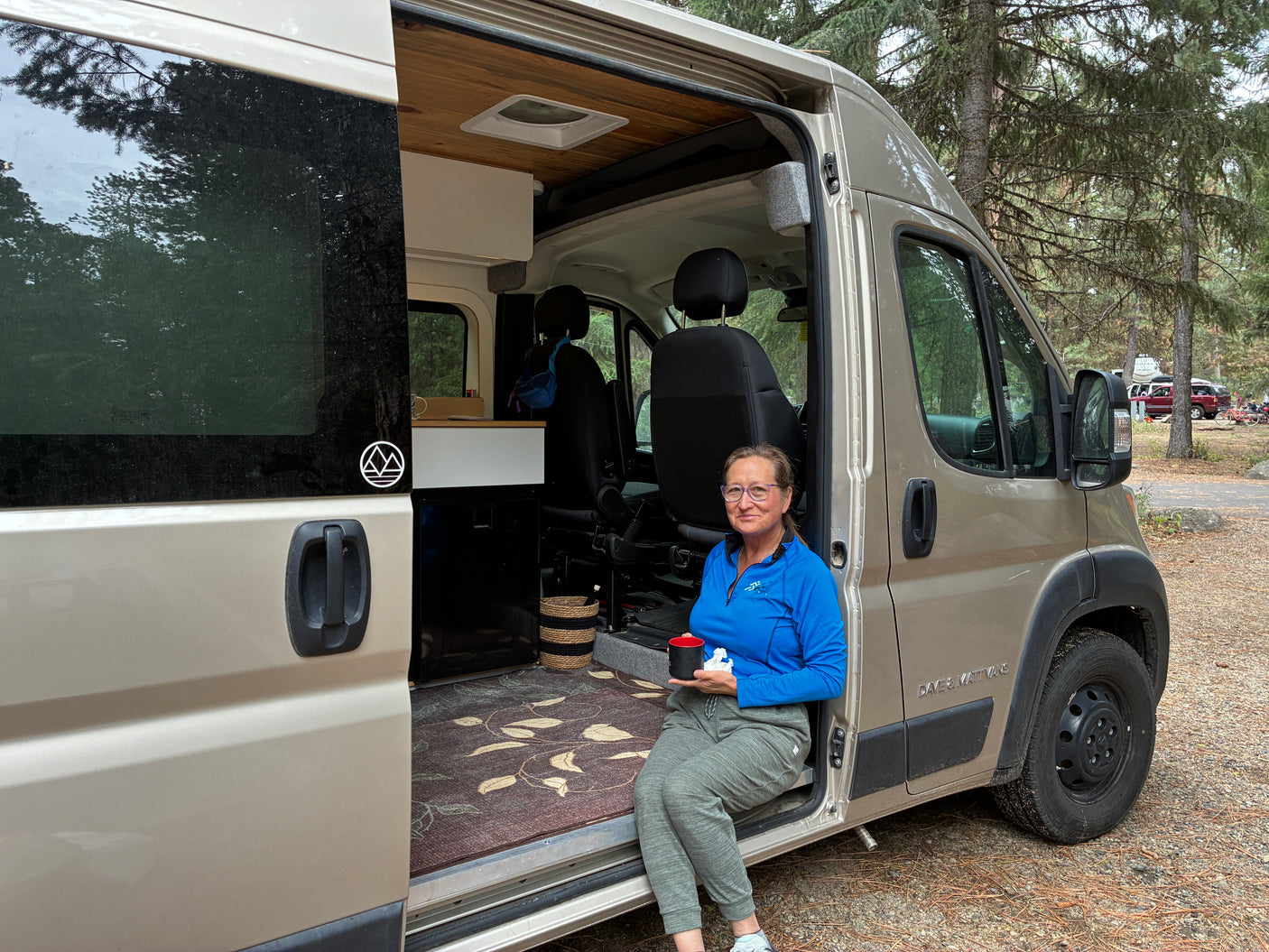 Enjoying a cozy moment in an **rv rental** camper van. A woman sits in the open doorway with a mug, surrounded by nature.