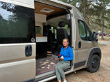 Enjoying a cozy moment in an **rv rental** camper van. A woman sits in the open doorway with a mug, surrounded by nature.