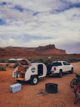 Teardrop RV rental for desert camping. White trailer with open kitchen, truck, and red rock landscape.