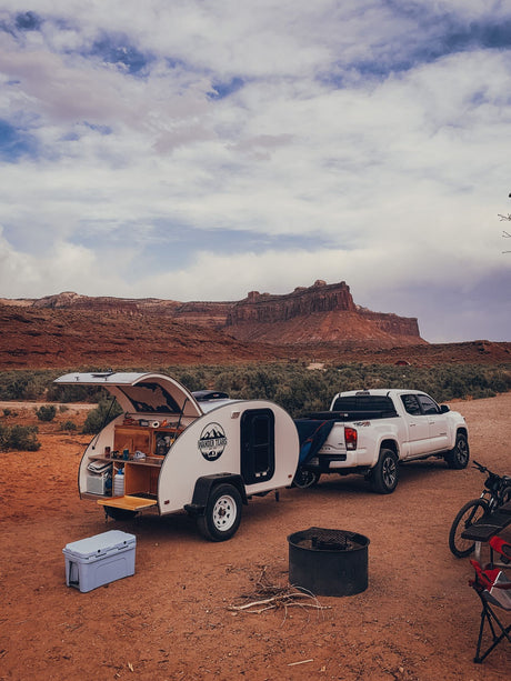 Teardrop RV rental for desert camping. White trailer with open kitchen, truck, and red rock landscape.
