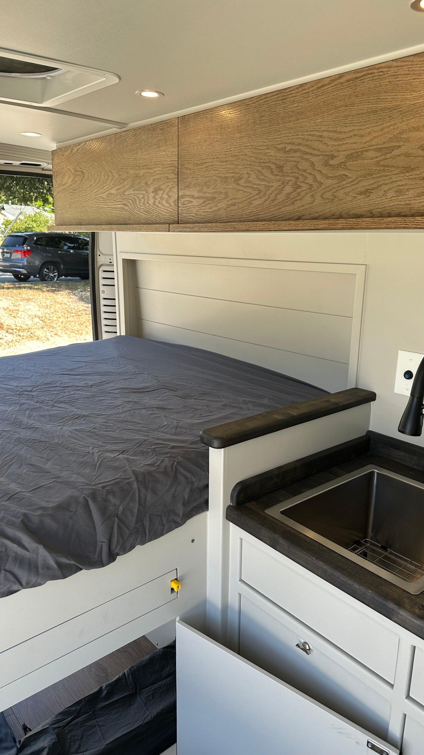 Interior of an rv rental: a grey bed on left, light wood cabinets above, and a compact kitchen sink on the right.
