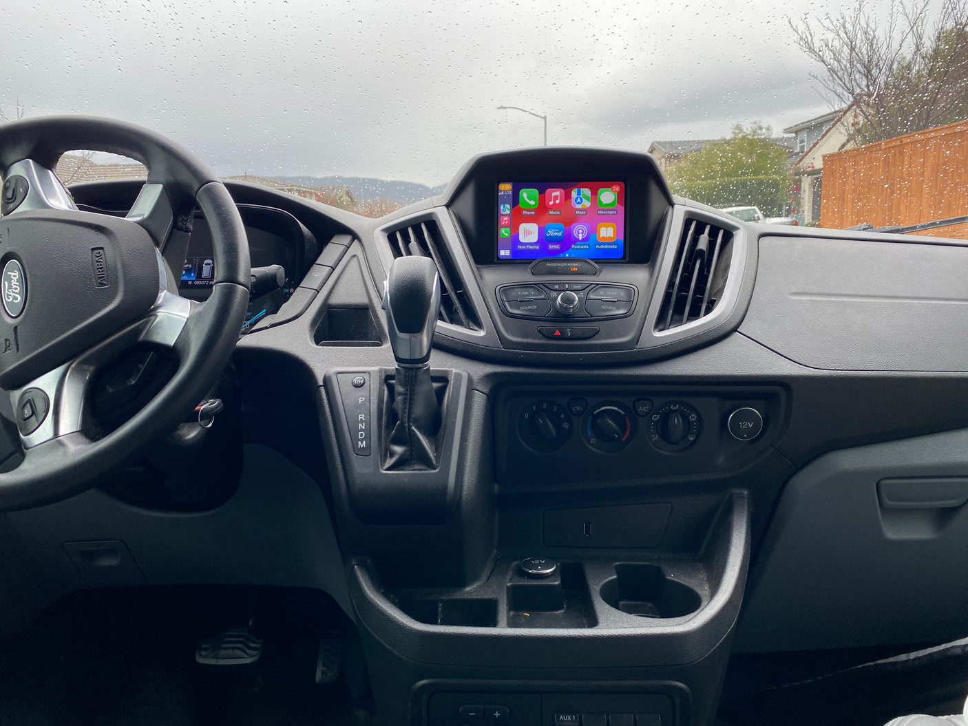 Inside a Ford Transit **rv rental** van, showing the dashboard, steering wheel, and infotainment screen with Apple CarPlay. Raindrops on windshield.