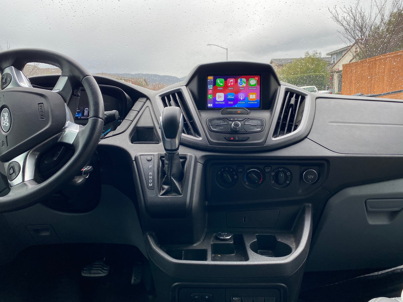 Inside a Ford Transit **rv rental** van, showing the dashboard, steering wheel, and infotainment screen with Apple CarPlay. Raindrops on windshield. — photo 36