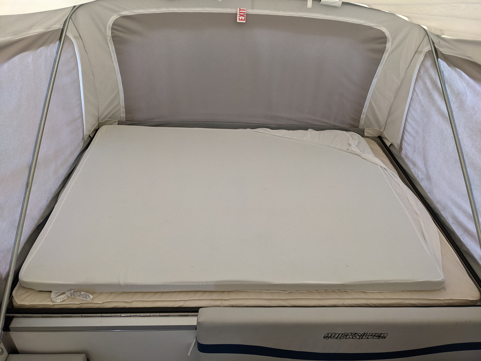 RV rental pop-up camper's interior bed: layered mattresses with an "EXIT" sign above. Quicksilver branding visible.