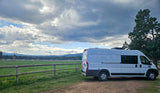 White RV rental camper van parked by a fence, offering views of a green field and majestic snow-capped mountains under cloudy skies.