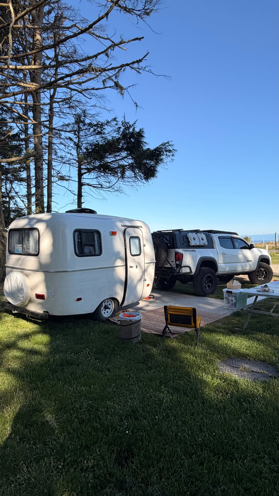 Cozy white travel trailer (rv rental) and adventure-ready pickup truck parked at a sunny campsite with trees.|A white — photo 1