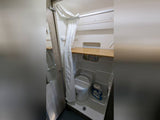 This compact RV rental bathroom features a white shower curtain, toilet, and wooden shelf, designed for efficient use of space.