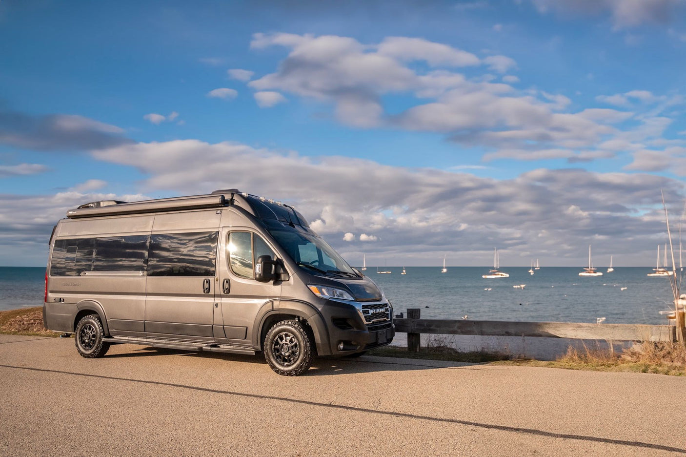 This sleek grey RV rental, a Class B camper van, is parked lakeside with sailboats in the background. Ready for your next adventure.