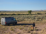 An RV rental camper van parked in a vast desert landscape with train tracks, a person, and their dog enjoying the open space.