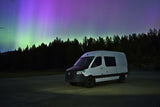 An RV rental camper van parked under stunning green and purple Northern Lights & starry sky in a forest.