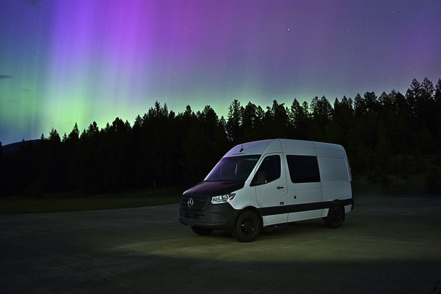 An RV rental camper van parked under stunning green and purple Northern Lights & starry sky in a forest.
