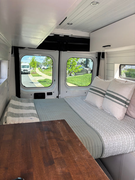 Cozy RV rental interior: bed with pillows, wooden table, and street views through large windows, perfect for travel.