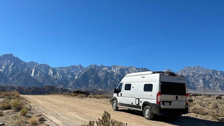 Experience mountain adventure with this RV rental. White camper van on a dirt road, framed by rugged mountains & clear sky.