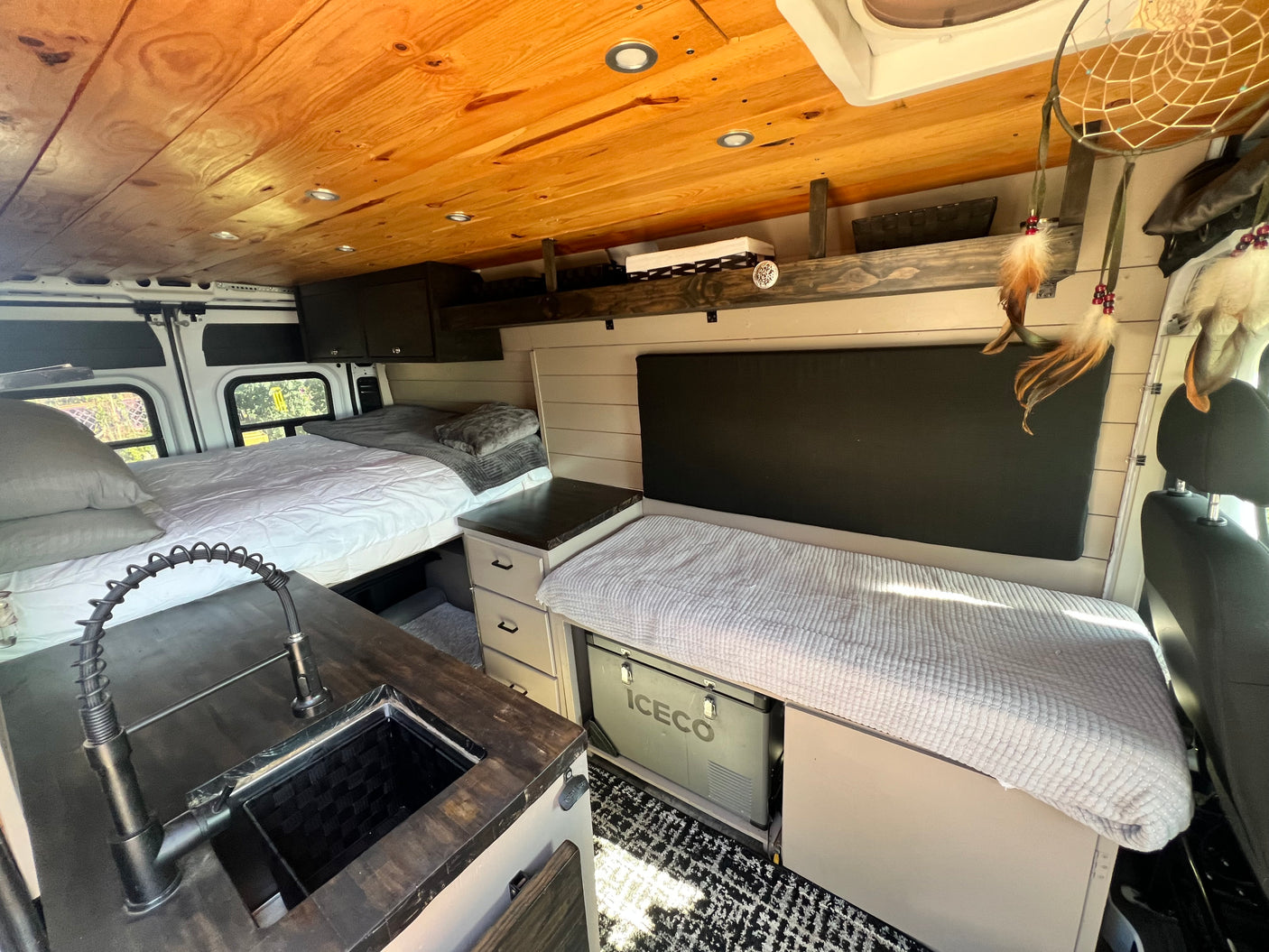 Inside a cozy rv rental camper van: A bed, sink with black faucet, light cabinetry, a bench over an ICECO fridge, and wood ceiling.