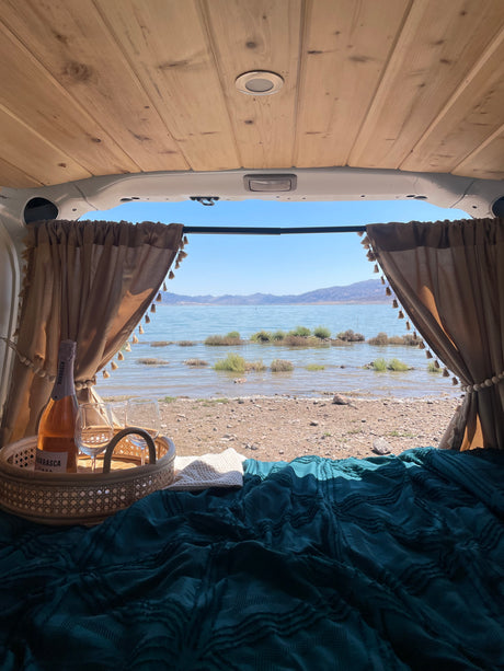 Enjoying a serene lake view from an RV rental, featuring a cozy bed, pulled-back curtains, and sparkling wine on a rattan tray.
