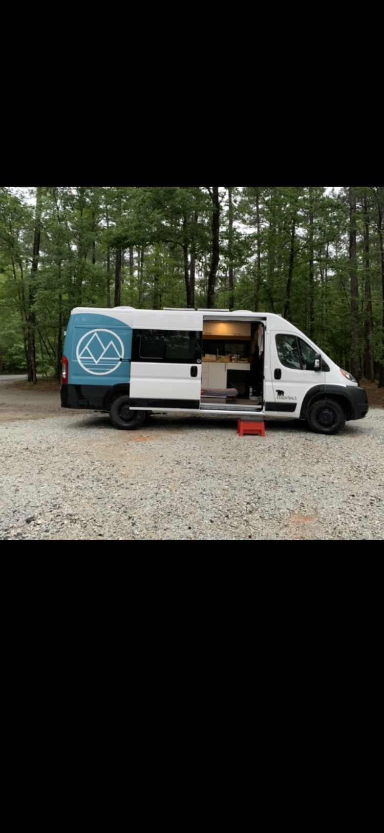 This white & blue custom camper van rv rental sits on gravel, forest background. Side door open, red step shows the cozy interior.