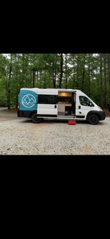 This white & blue custom camper van rv rental sits on gravel, forest background. Side door open, red step shows the cozy interior.
