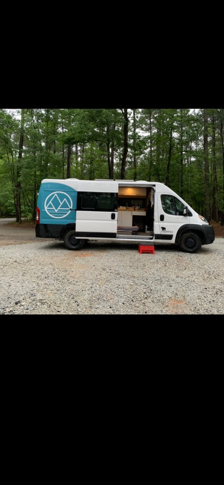 This white & blue custom camper van rv rental sits on gravel, forest background. Side door open, red step shows the cozy interior.
