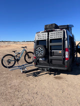 RV rental: Dark grey camper van with roof AC & bike rack, holding a light mountain bike, parked in sandy terrain.
