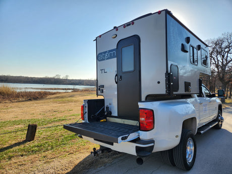 White dually pickup truck with a mounted Atom TL RV rental truck camper parked by a lake under a clear sky.